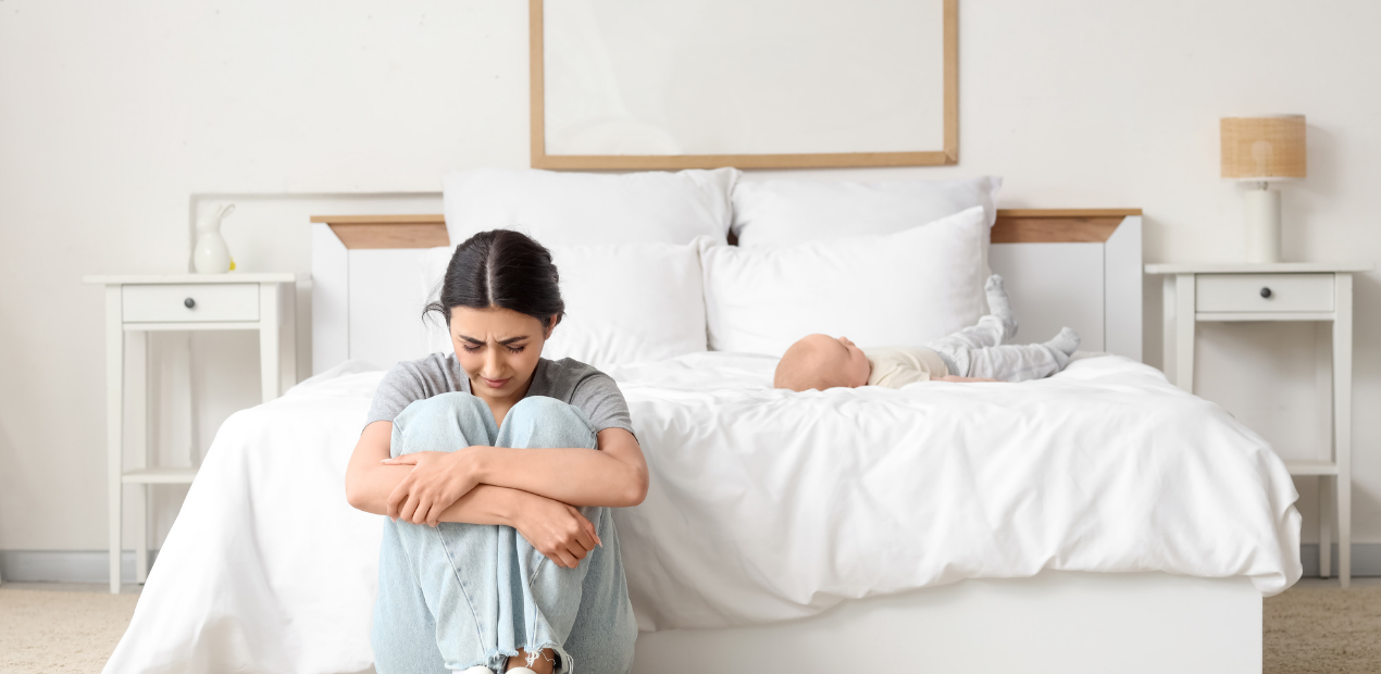 Fotografia de uma mãe sentada no chão do quarto com expressão de angústia, enquanto o bebê descansa na cama ao fundo, ilustrando o isolamento emocional típico dos sintomas da depressão pós parto.
