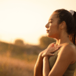 Mulher em ambiente natural ao pôr do sol, com olhos fechados e mãos sobre o peito, realizando uma prática de respiração consciente ou mindfulness, que são exemplos de exercícios para ansiedade.