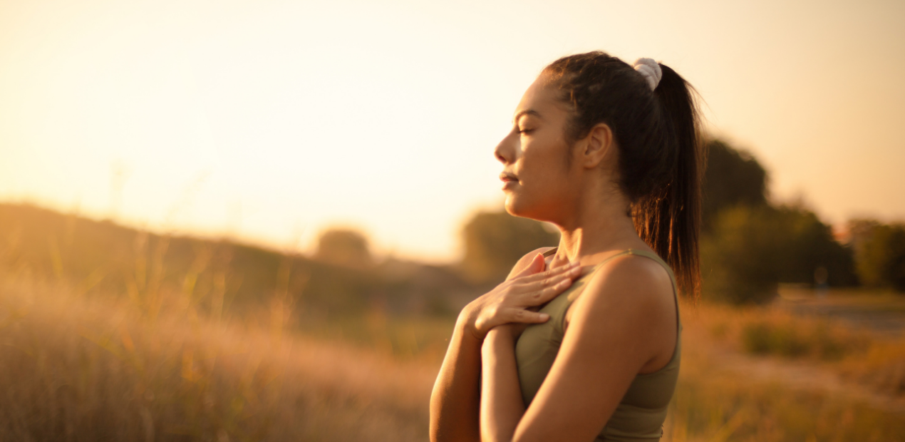 Mulher em ambiente natural ao pôr do sol, com olhos fechados e mãos sobre o peito, realizando uma prática de respiração consciente ou mindfulness, que são exemplos de exercícios para ansiedade.
