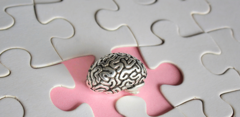 Fotografia de uma pequena escultura de cérebro prateado sobre uma peça de quebra-cabeça rosa central, rodeada por peças brancas, simbolizando o desafio de compreender a complexidade neurobiológica dos sintomas de esquizofrenia.