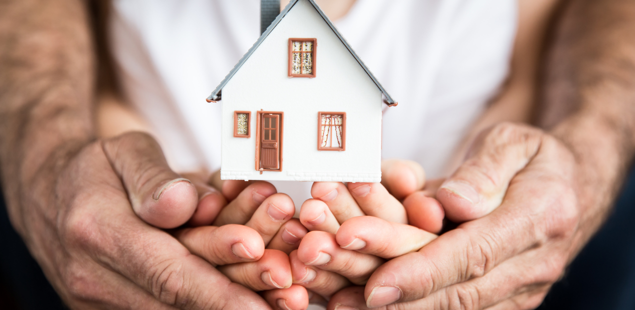 Mãos de adultos e crianças segurando uma miniatura de casa, simbolizando proteção, segurança emocional e bem-estar familiar