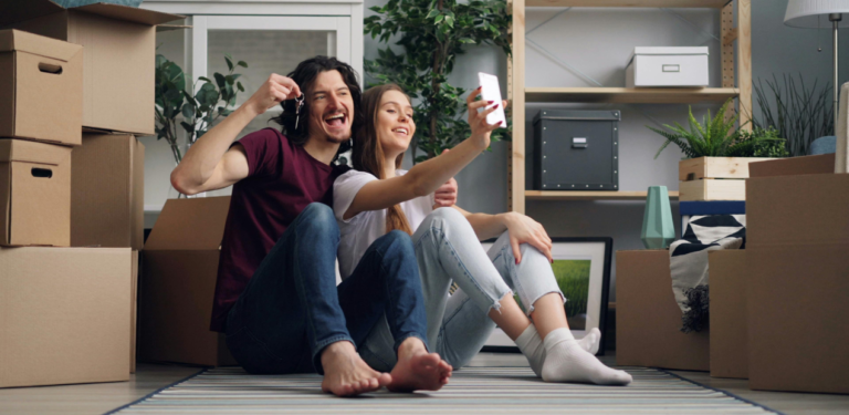 Casal sentado no chão entre caixas de mudança, sorrindo em um novo lar, simbolizando renovação emocional e bem-estar familiar