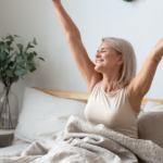 Mulher acordando em um quarto iluminado, alongando os braços, simbolizando bem-estar mental e renovação em um ambiente acolhedor