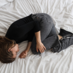 Pessoa deitada na cama em posição fetal, com corpo encolhido e expressão neutra, sugerindo cansaço emocional