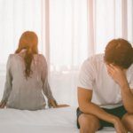 Homem sentado na cama com a mão no rosto em sinal de angústia, enquanto a mulher está de costas, indicando distanciamento no relacionamento