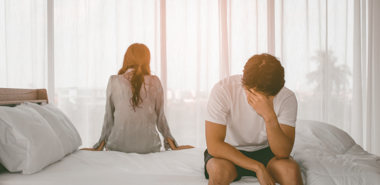 Homem sentado na cama com a mão no rosto em sinal de angústia, enquanto a mulher está de costas, indicando distanciamento no relacionamento