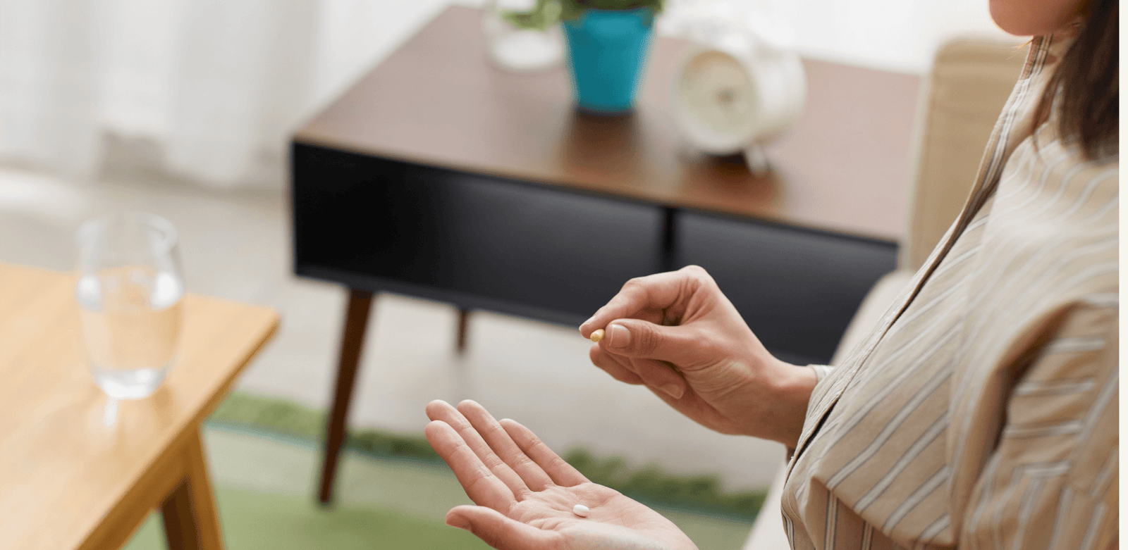 Pessoa sentada segura um comprimido próximo à mão aberta, em ambiente doméstico com copo de água sobre a mesa