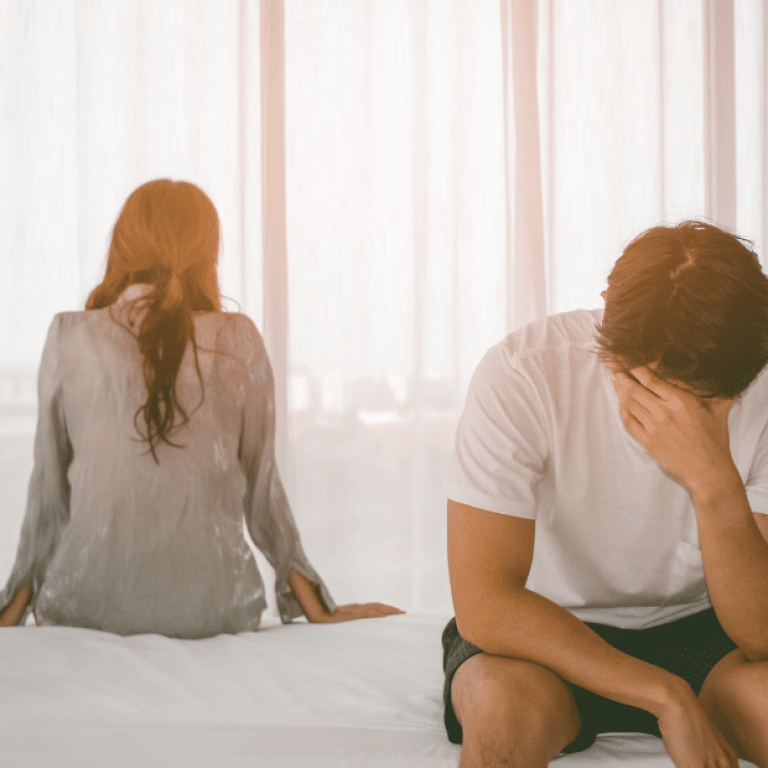 Homem sentado na cama com a mão no rosto em sinal de angústia, enquanto a mulher está de costas, indicando distanciamento no relacionamento