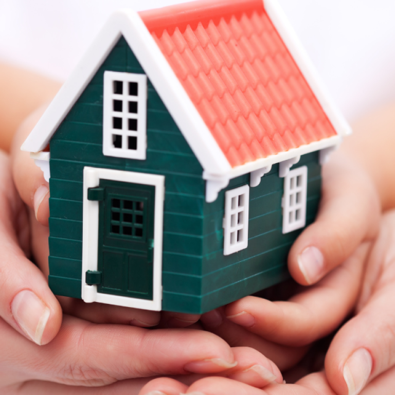 Casa em miniatura sendo segurada por duas mãos, representando segurança, acolhimento e a busca por um lar que vai além do status social
